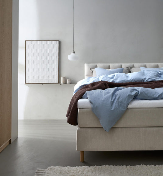 Lyst soveværelse med beige kontinentalseng redt med lyseblåt sengetøj og brun plaid, hvid pendel og billede på hylde.