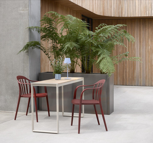 To røde udestole ved et smalt bord med lille bærbar lampe og plante, foran en moderne facade med store bregner.