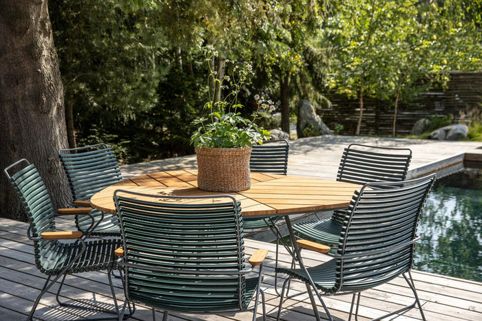 Rundt teakbord med grønne metalstole på en træterrasse ved en pool, med en fletkurv med plante i midten i et solrigt havemiljø.