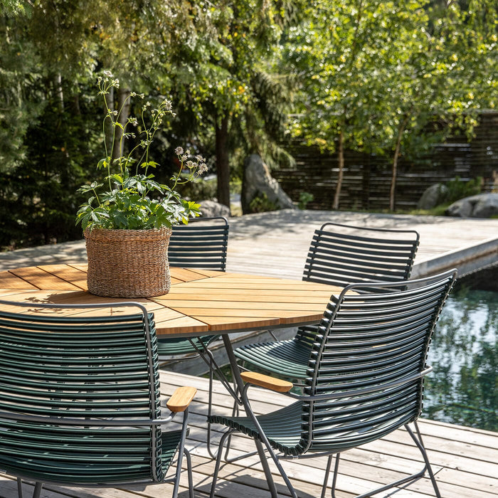 Rundt teakbord med grønne metalstole på en træterrasse ved en pool, med en fletkurv med plante i midten i et solrigt havemiljø.