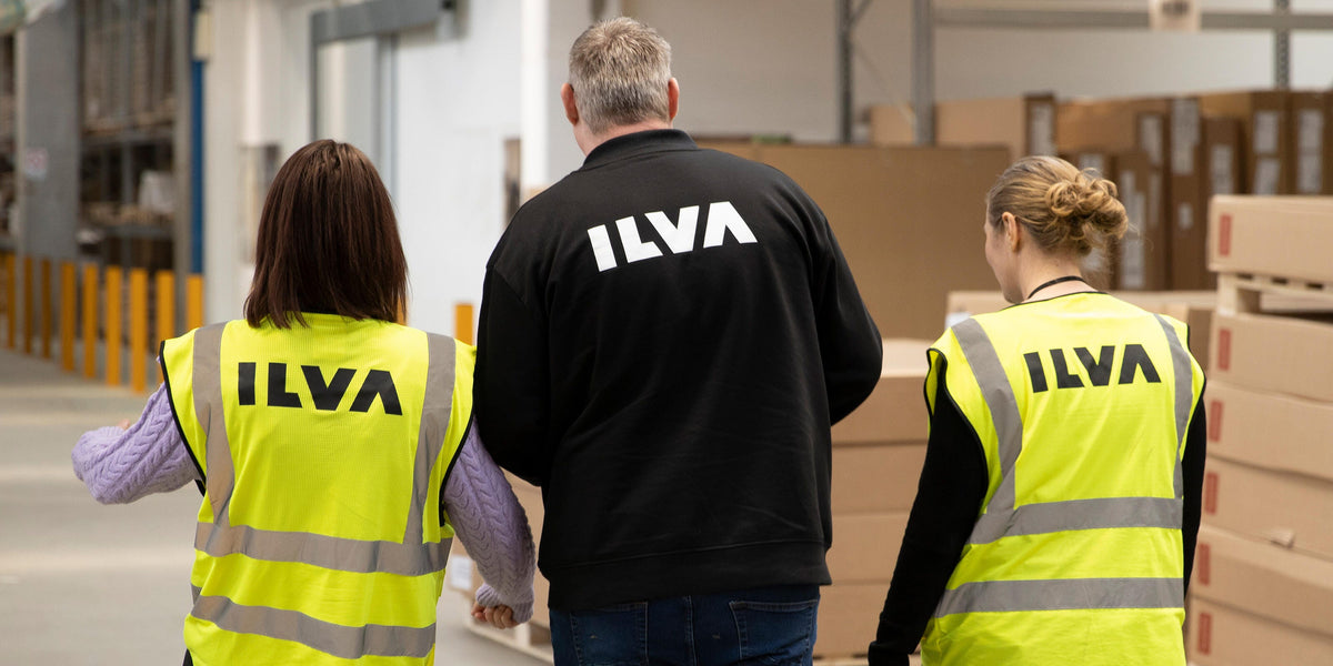 Tre personer med ILVA‑logo på sikkerhedsveste og jakke går gennem et lager med stablede kasser.