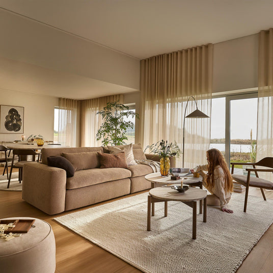 Lyst og varmt opholdsrum med beige sofa, fuldlange, lette gardiner og to runde sofaborde; en person sidder på tæppet med en kop og en bog.