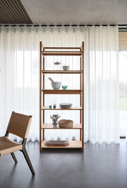 Åben reol i lyst træ med keramikskåle, glas og fletkurv foran lyse transparente gardiner; fletstol i venstre side.