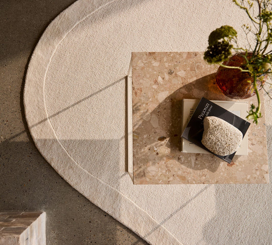 Set oppefra: terrazzo-bord på lyst, buet tæppe; ravfarvet vase, bog med titlen ”Recognize” og lys sten i varmt dagslys.