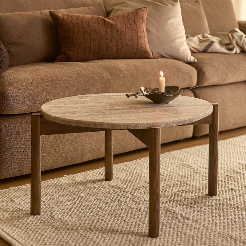 Rundt sofabord i træ på beige tæppe foran brun sofa med puder og en tændt lysdekoration.