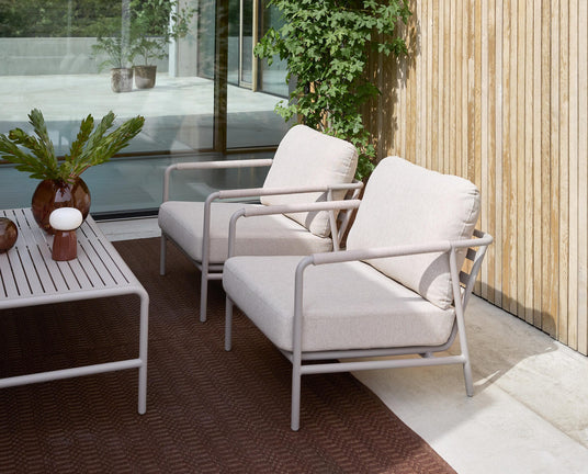 Terrasse med to beige loungestole med rørformet stel, ribbet sofabord med vaser og grønne kviste, brun udendørs måtte ved træbeklædt væg og glasdør i dagslys.