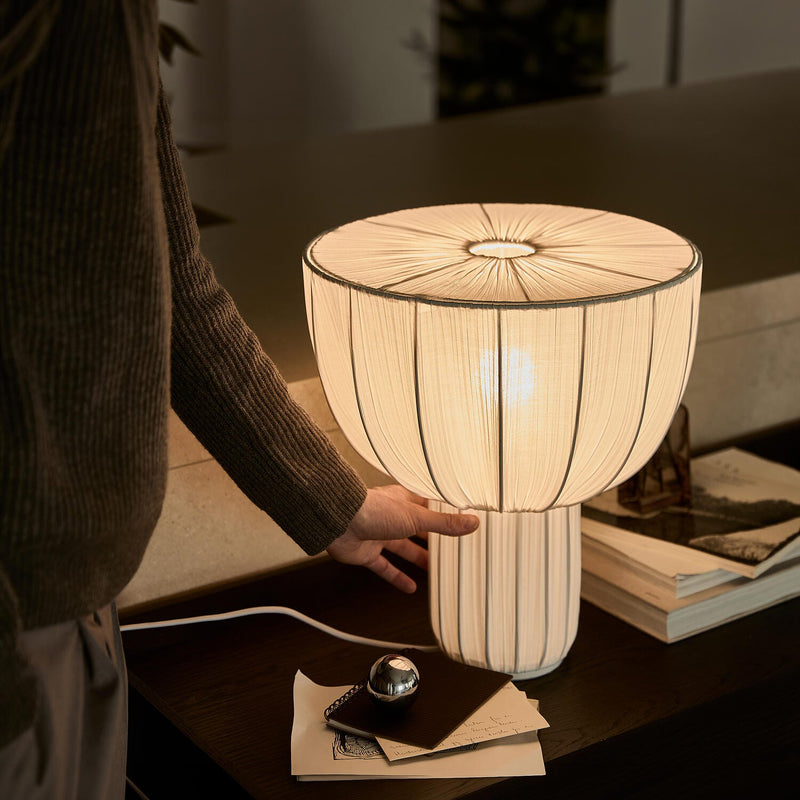 Hånd der justerer en varmglødende bordlampe med plisseret skærm på et mørkt sidebord med bøger.