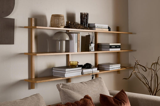 Vægmonteret reol i lyst træ over beige sofa med puder; bøger som Chanel og New Nordic Houses, lille bordlampe og flettet kurv på hylderne.