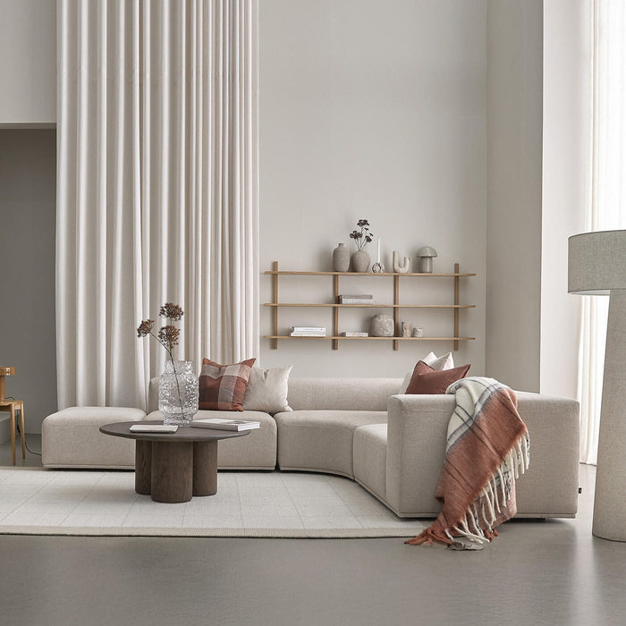 Beige modulsofa med terrakottapuder og plaid i en minimalistisk stue, rundt sofabord i mørkt træ, gulvlampe og væghylde med vaser og bøger; lyse gardiner og neutrale toner.