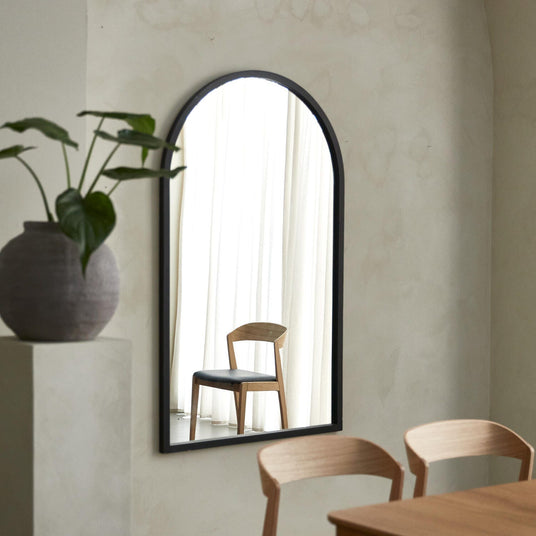 Bueformet vægspejl med sort ram; spejlet reflekterer en træstol med sort sæde foran lyse gardiner. I forgrunden ses træstole, et bord og en vase med grøn plante i et lyst, minimalistisk rum.