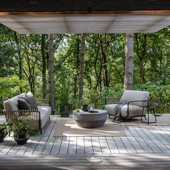 Terrasse på trædæk i skov med loungesofa og lænestol, rundt betonbord, tæpper, krukplanter og stablet brænde under pergola.