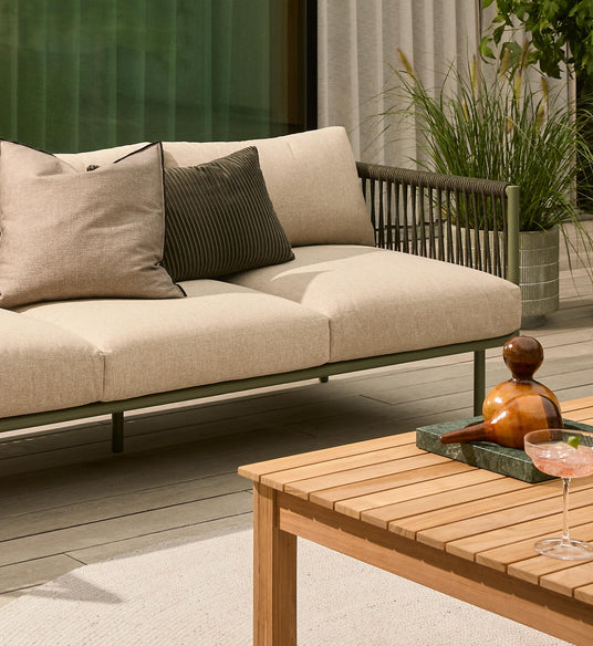 Beige udendørs sofa med puder ved siden af et lameltræbord på en moderne terrasse, med grønne planter og lyse gardiner i baggrunden.