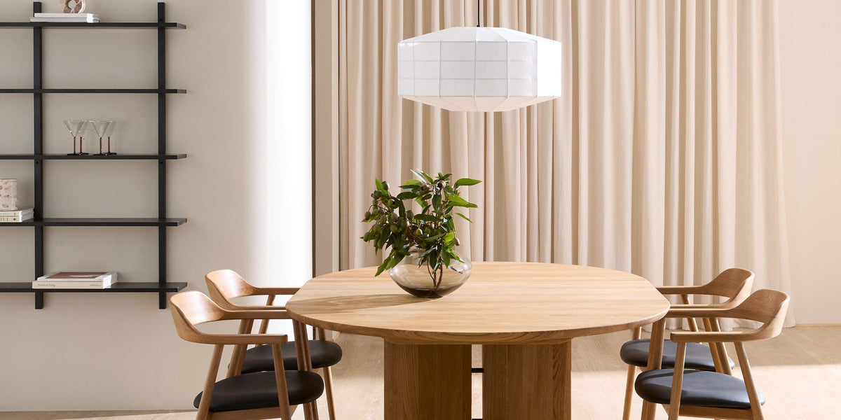 Minimalistisk spisestue med rundt egetræsbord og fire stole, hvid pendellampe, beige gardiner og sort væghylde; en glasvase med grønne grene står på bordet.