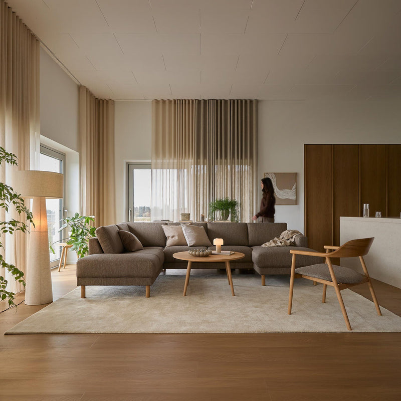 Lys skandinavisk stue med beige hjørnesofa, træfåtølj og sofabord på lys tæppe, lette gardiner, grønne planter og et tændt lys.