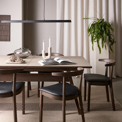 Minimalistisk spisestue med mørke træstole, beige bord, pendellampe, tændte lys, glasvase, åben bog og høj grøn plante foran lyse gardiner.