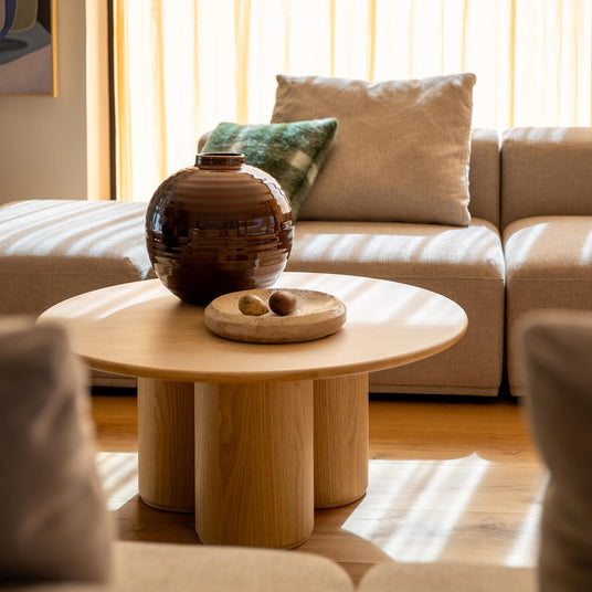 Beige sofa og rundt sofabord i lyst træ med brun keramikvase og skål, solstriber gennem lyse gardiner.