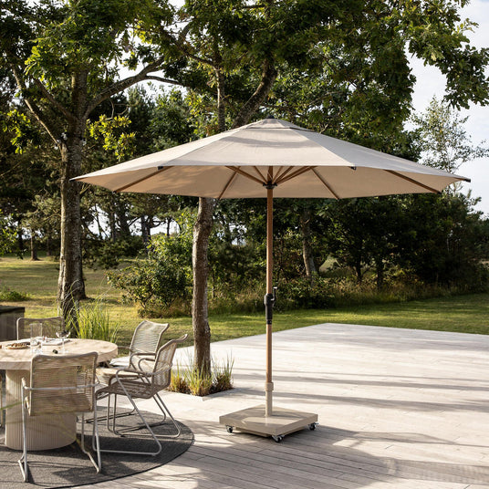 Beige parasol på rullefod på terrasse ved siden af rundt spisebord med trådstole; have og træer i baggrunden i dagslys.