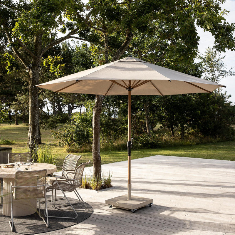 Beige parasol på rullefod på terrasse ved siden af rundt spisebord med trådstole; have og træer i baggrunden i dagslys.