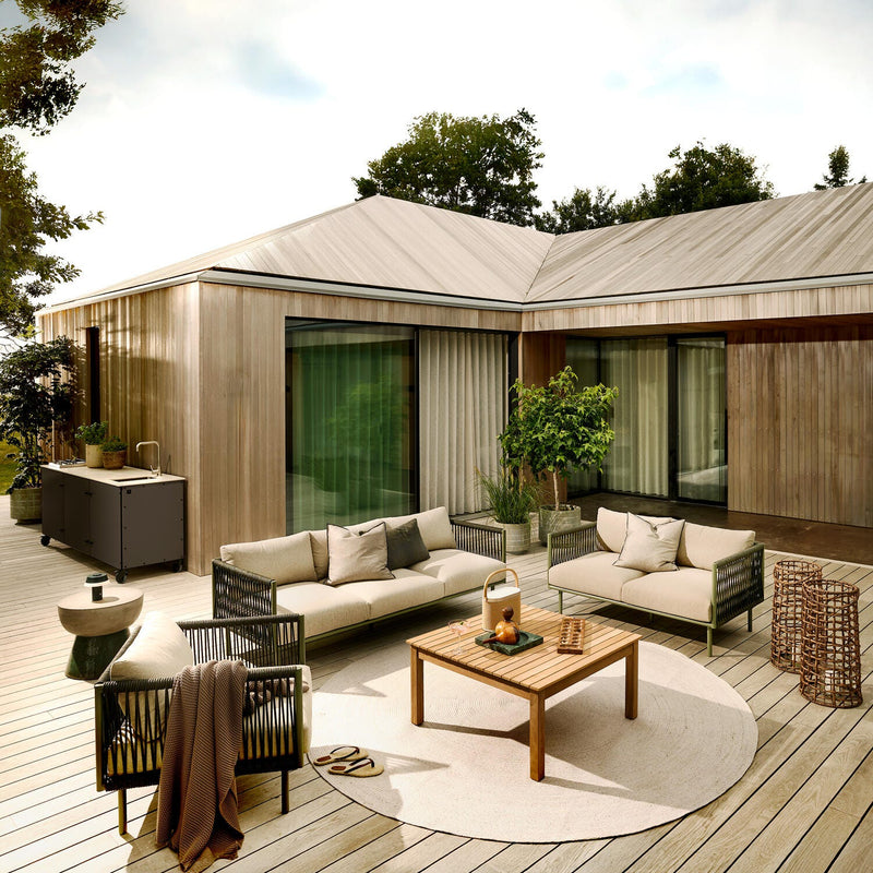 Moderne træterrasse med lyse loungesofaer, træbord, rundt tæppe, lanterne og potteplanter foran et hus med skydedøre.