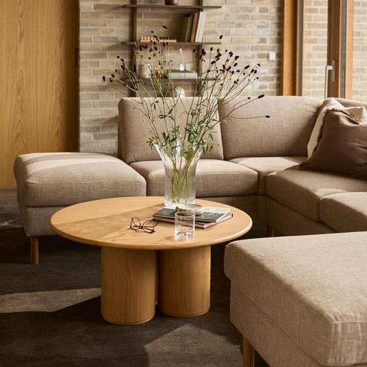Beige hjørnesofa i stue med rundt sofabord i træ, glasvase med fine blomster, bøger og et vandglas.