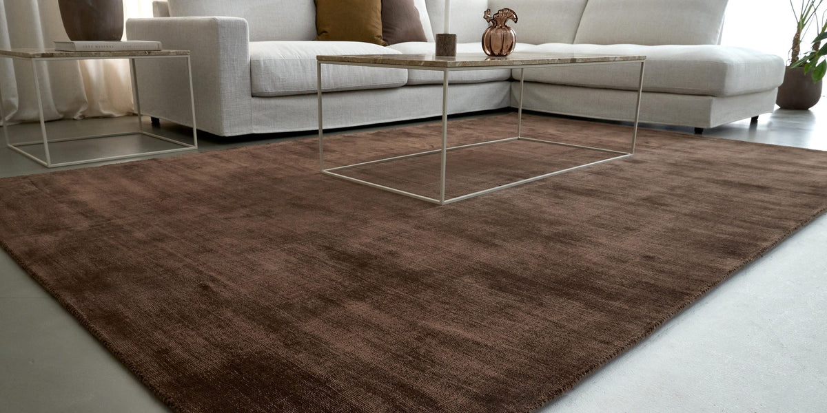 Stue med et stort brunt rektangulært tæppe, hvid hjørnesofa, slankt metalsofabord med marmortop, puder i brun og okker, sidebord med bog og vase, lette gardiner og dagslys.