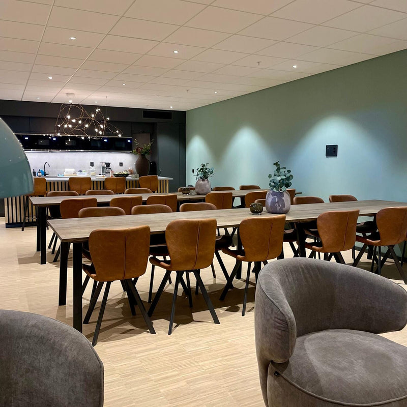 Stor spisestue med to lange træborde og brune læderstole, grøn væg og åbent køkken i baggrunden med moderne pendellampe og planter.