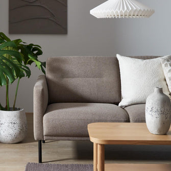 Beige sofa med hvide puder, lyst sofabord i træ, keramikvase, hvid plisseret pendel og grøn monstera i en stue.