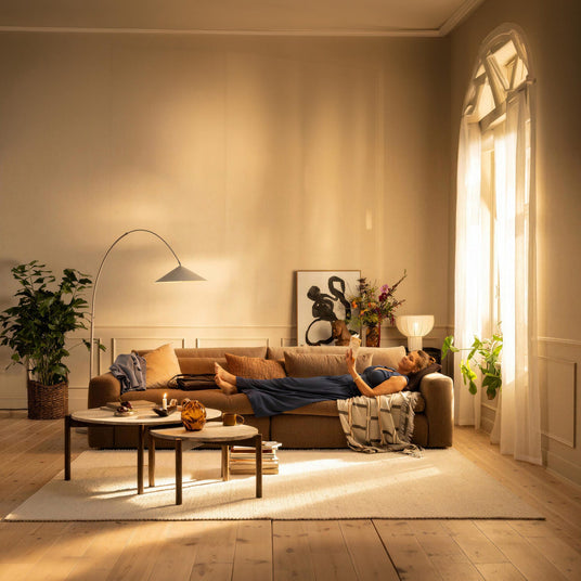 Stue i varmt dagslys med kvinde der læser på brun sofa, runde sofaborde, buet gulvlampe, planter og lyse gardiner.