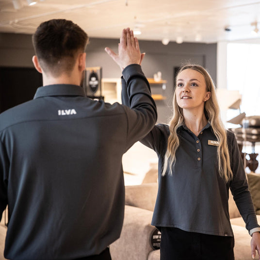 To ILVA‑medarbejdere i mørke uniformer giver hinanden en high‑five i et møbelshowroom.