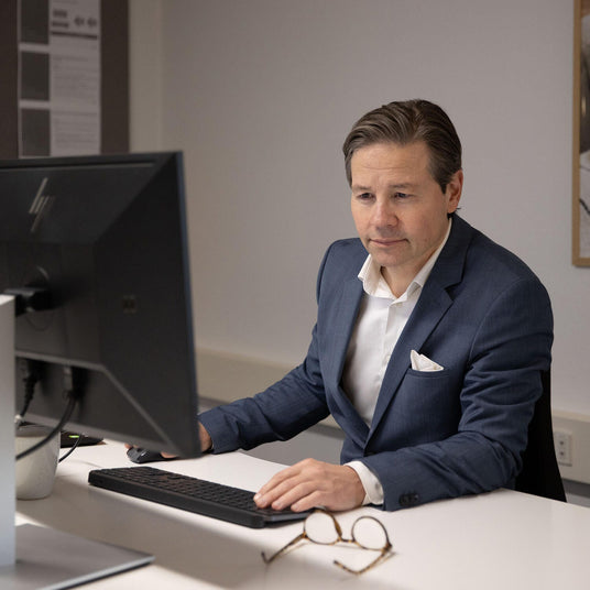 Person i jakkesæt arbejder ved to computerskærme på et kontor, hænder på tastatur og mus; briller ligger på skrivebordet.