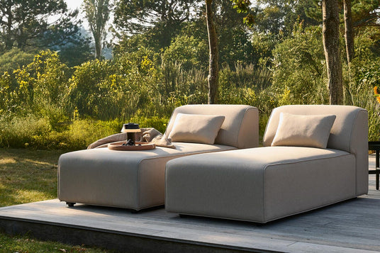To beige loungemoduler med puder på en træterrasse i en grøn have i dagslys.