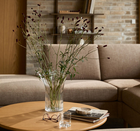 Vase med lange grønne grene på rundt sofabord i stue med beige hjørnesofa; bøger, briller og et vandglas ved siden af.