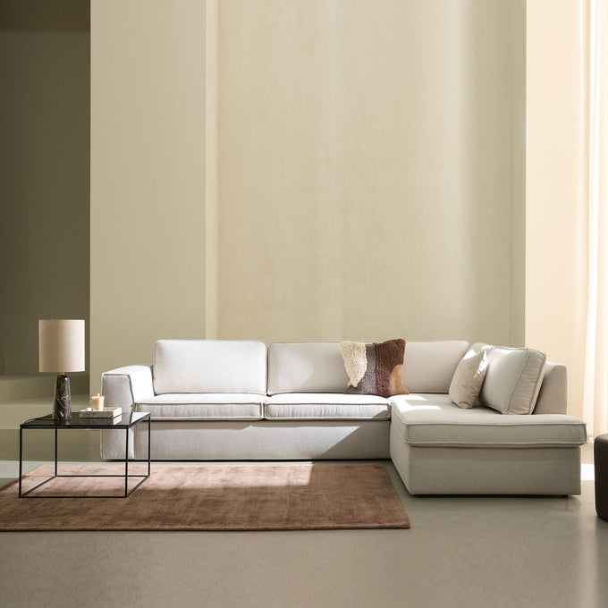 Lyst, minimalistisk stue med beige modulsofa med chaiselong, glasbord med bordlampe og lys, brun tæppe og blødt dagslys gennem lyse gardiner.
