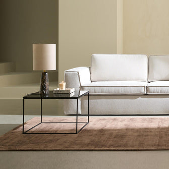 Lyst, minimalistisk stue med beige modulsofa med chaiselong, glasbord med bordlampe og lys, brun tæppe og blødt dagslys gennem lyse gardiner.