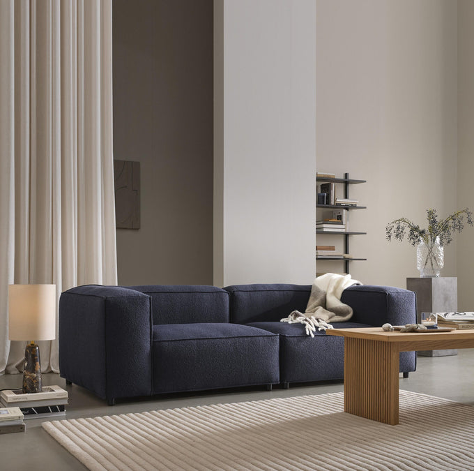 Mørkeblå modulsofa i stue med lyse gardiner, rillet sofabord i træ, beige ribbet tæppe, bordlampe, hylde med bøger og glasvase med grene; plaid over sofaen.
