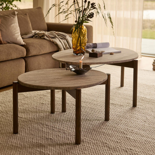 To runde sofaborde i træ på et lyst struktur-tæppe, med ravfarvet vase med grene og et tændt lys; beige sofa og lette gardiner i dagslys.