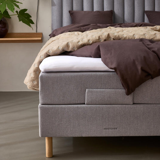 Grå polstret seng med lodret rillet sengegærde, brunt sengetøj og beige quilt, træben og mærket nocturne; ved siden af en væghylde med vase, grøn plante og væglampe.