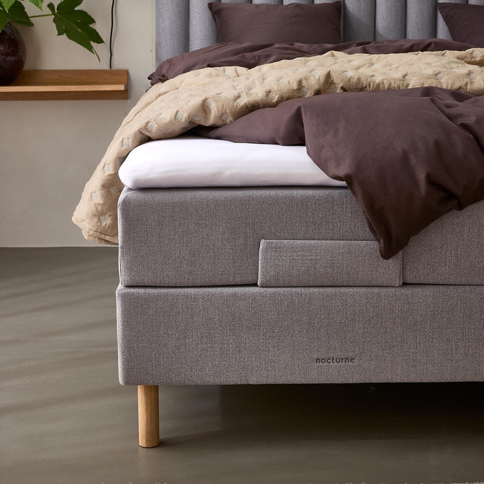 Grå polstret seng med lodret rillet sengegærde, brunt sengetøj og beige quilt, træben og mærket nocturne; ved siden af en væghylde med vase, grøn plante og væglampe.