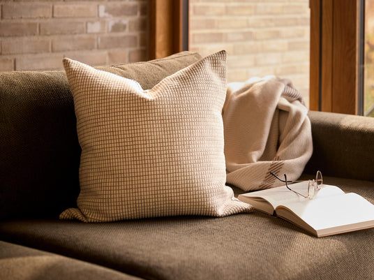 Beige pude med rutemønster på mørk sofa, med plaid, opslået bog og briller ved et vindue i varmt dagslys.
