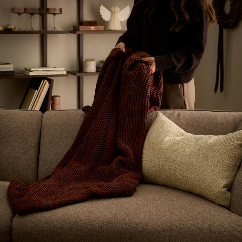 Person lægger en rustbrun strikplaid over en beige sofa med lys pude i en hyggelig stue.