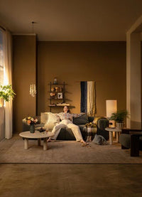 Stue med varm brun væg, grøn sofa og en person der slapper af; runde sofaborde med blomster i vase, plaid og puder, gulvlampe, hængependel og store gardiner ved vinduet.