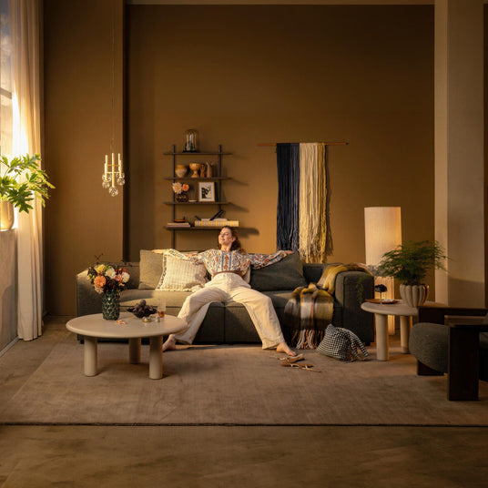 Stue med varm brun væg, grøn sofa og en person der slapper af; runde sofaborde med blomster i vase, plaid og puder, gulvlampe, hængependel og store gardiner ved vinduet.