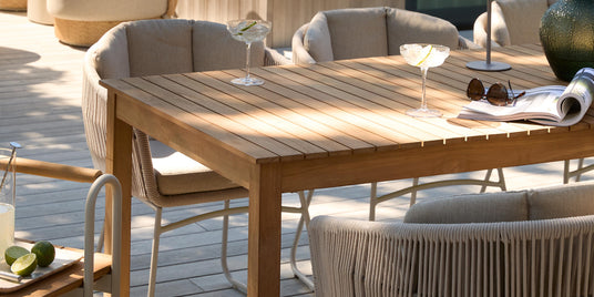 Solrig terrasse med spisebord i træ og beige snore-stole; to cocktailglas, lille bordlampe, grøn vase og opslået magasin på bordet.