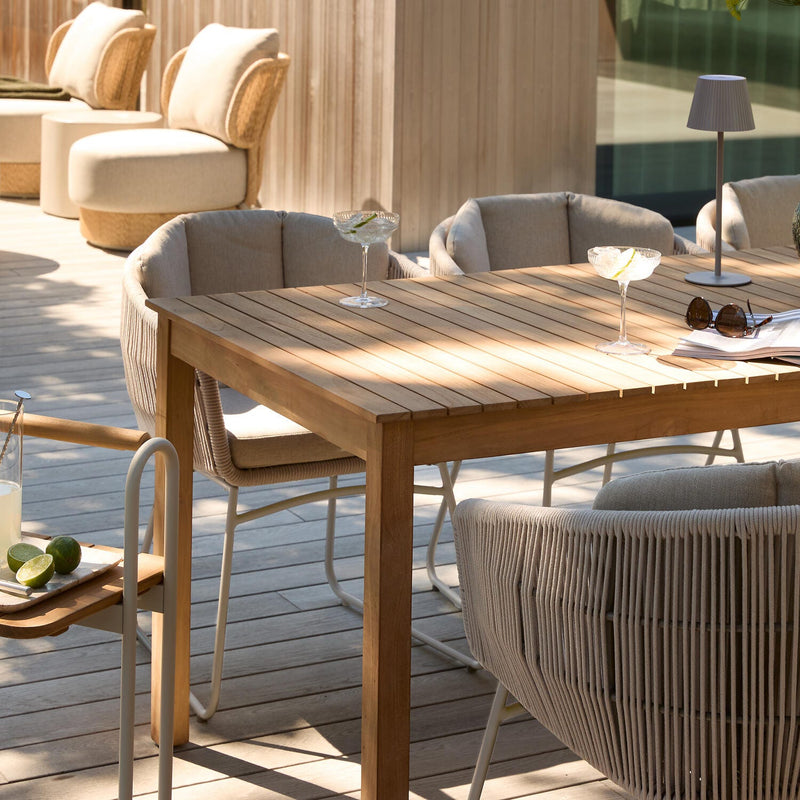 Solrig terrasse med spisebord i træ og beige snore-stole; to cocktailglas, lille bordlampe, grøn vase og opslået magasin på bordet.