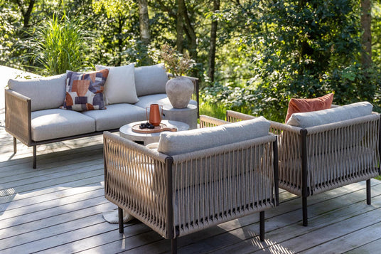 Lysegrå havemøbler på træterrasse i skov: sofa og to loungestole omkring rundt bord med kande og fad, puder og vase med grene i sollys.