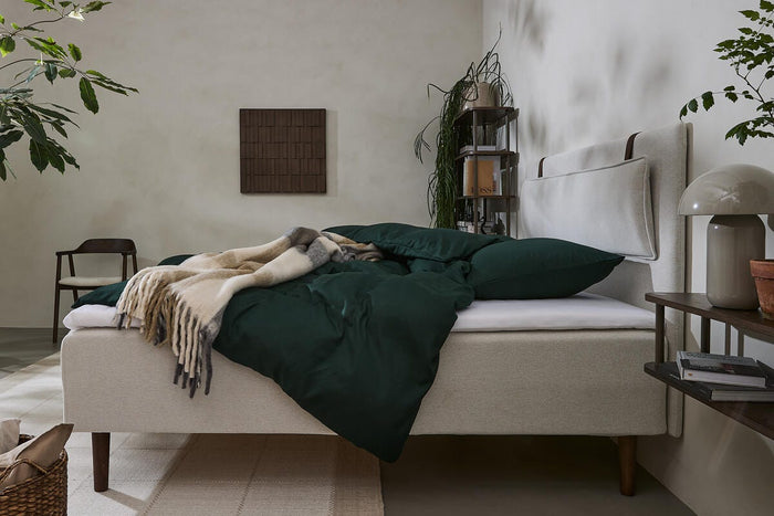 Lyst soveværelse med beige polstret seng, mørkegrønt sengetøj, uldtæppe og planter.