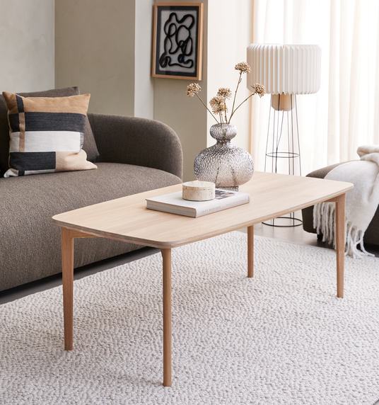 Lyst sofabord i eg på lys tekstureret tæppe med glasvase med tørrede blomster, bog og lille lys i et skandinavisk stue-miljø med sofa, gulvlampe og gardiner i dagslys.