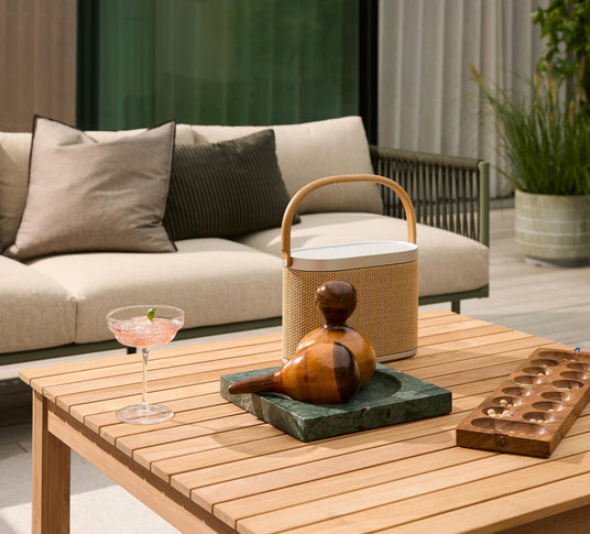 Udendørs terrasse med beige sofa og puder; på teakbordet står et glas lyserød drink med lime, en bærbar højttaler, en træskulptur på grøn stenplade og et mancala‑spil; rolig sommerstemning.