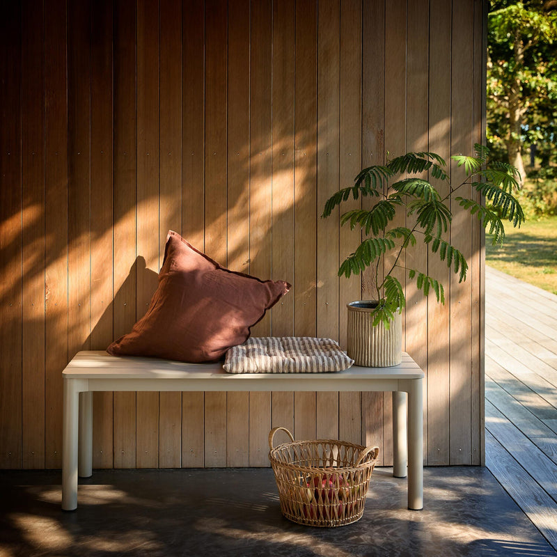 Lyst træbeklædt væg med bænk, brun pude, stribet hynde, potteplante og kurv i sollys ved terrasse.