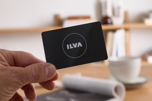 Hånd der holder et sort kort med ILVA‑logo, med sløret boligmiljø, kop og magasin på bordet i baggrunden.